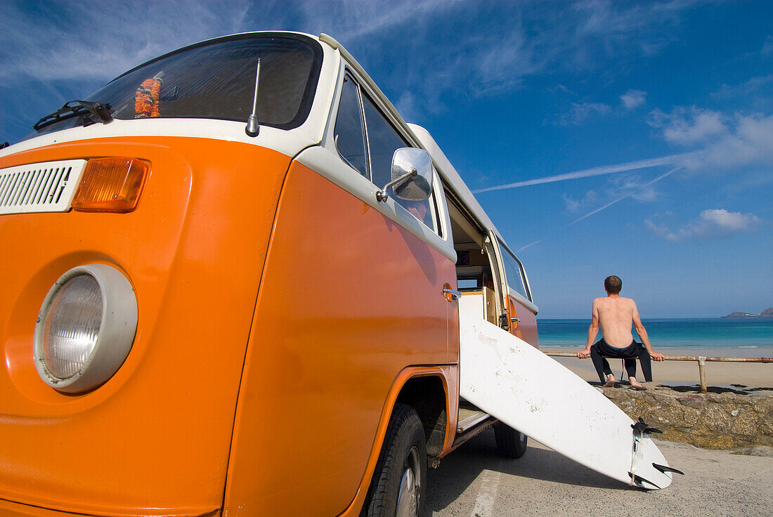 Surfer and board beside orange and white old campervan parked beside the sea, Sennen Cove, Cornwall, England