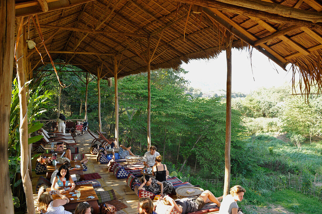 Travellers relaxing at reataurant terrace under palm roof, Nam Khan river, tributary to Mekong river, Luang Prabang, Laos