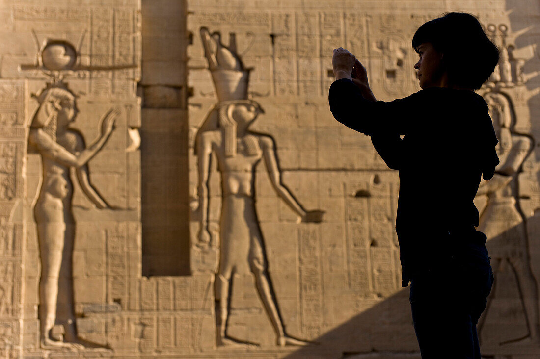 Silhouette of tourist taking photographs in front of large reliefs on walls of Second Pylon, Temple of Isis, Philae Island, near Aswan, Egypt