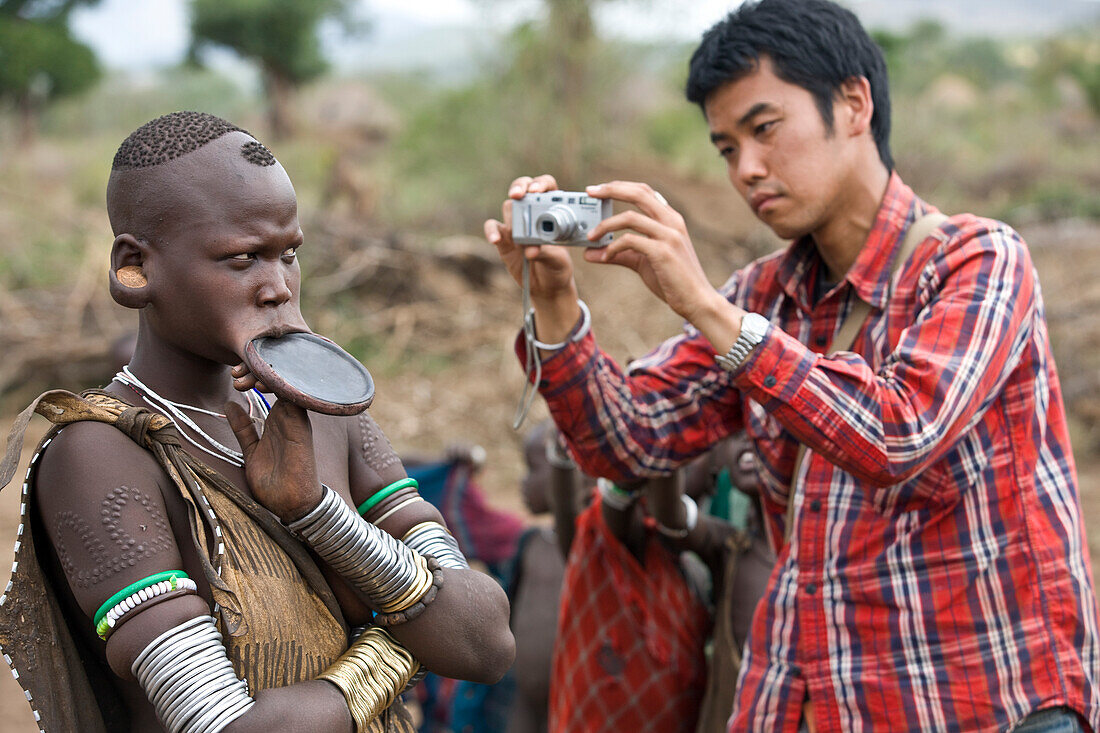 Japanese Tourist Taking A Picture Of A Slightly Annoyed Mursi Tribal Woman Wearing A Large Pottery Lip-Plate (Dhebi), Maridungka Village / Mursiland / South Omo / Southern Nations, Nationalities & People'S Region, Ethiopia