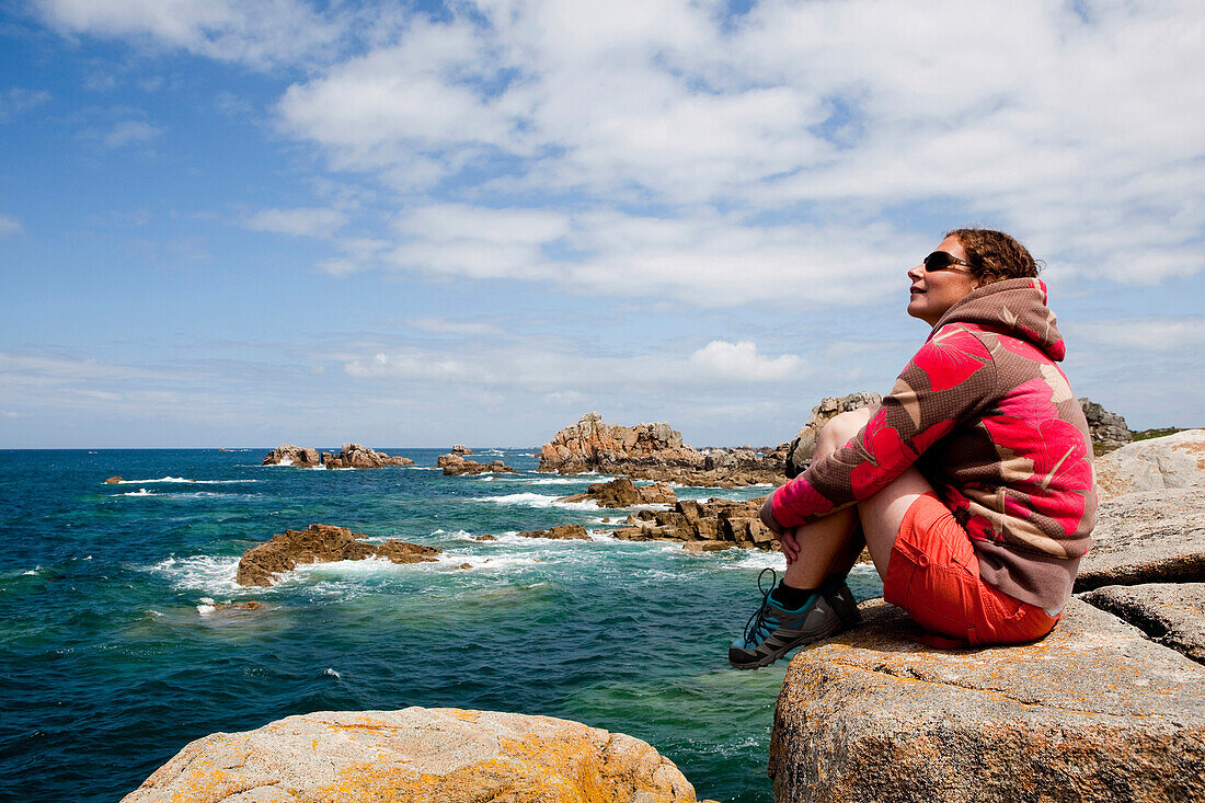 Young woman sitting looking out to sea on rocky shoreline in Brittany, Brittany, France.