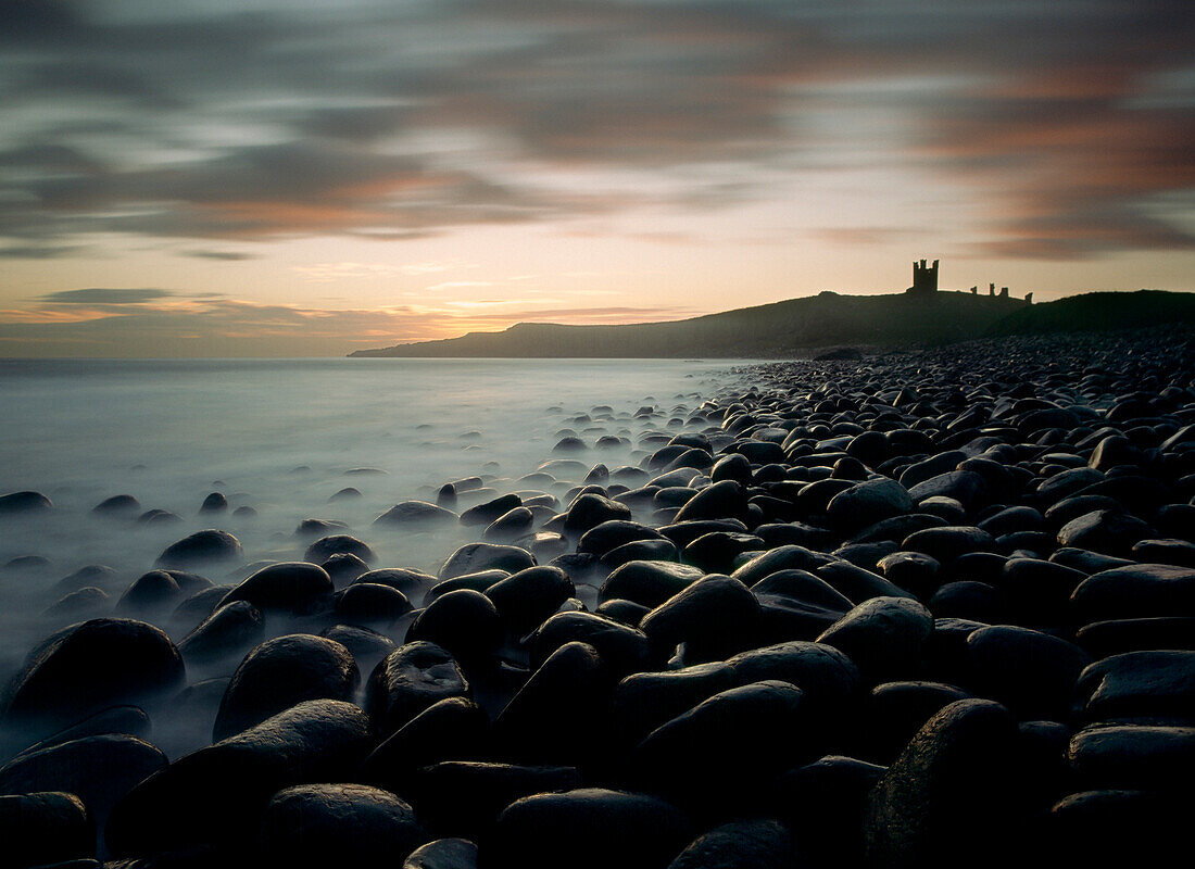 Looking along boulder covered beach towards Dunstanburgh Castle at dawn, Northumberland, England