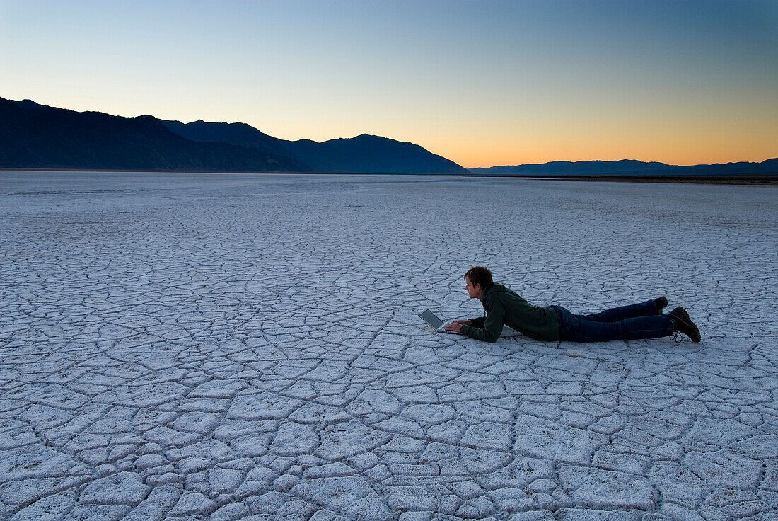 Man lying down using laptop on the salt pans at dawn, Death Valley, California