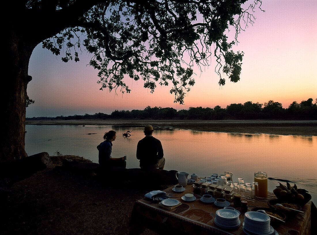 Couple sitting on log with hot drinks before dawn, South Luangwa National Park, Zambia