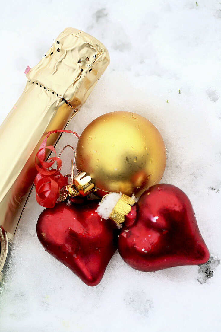 Two red satin glass hearts with champagne bottle neck and gold ball on snow