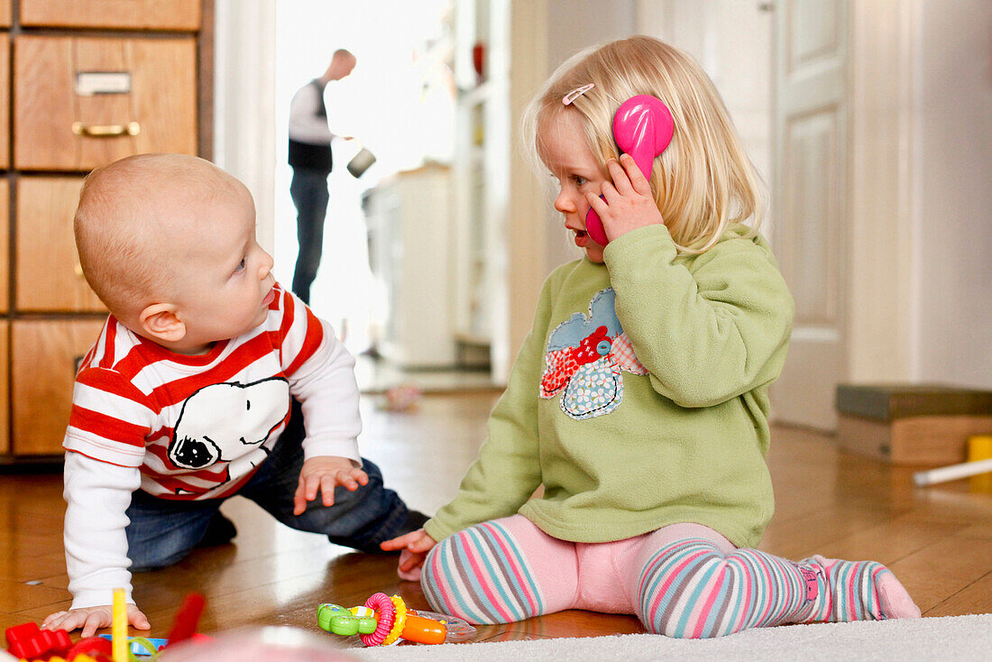 Kleiner Junge und kleines Mädchen spielen miteinander, Telefon, Vater im Hintergrund, Junge 9 Monate, Mädchen 2 Jahre, MR, Leipzig, Sachsen, Deutschland