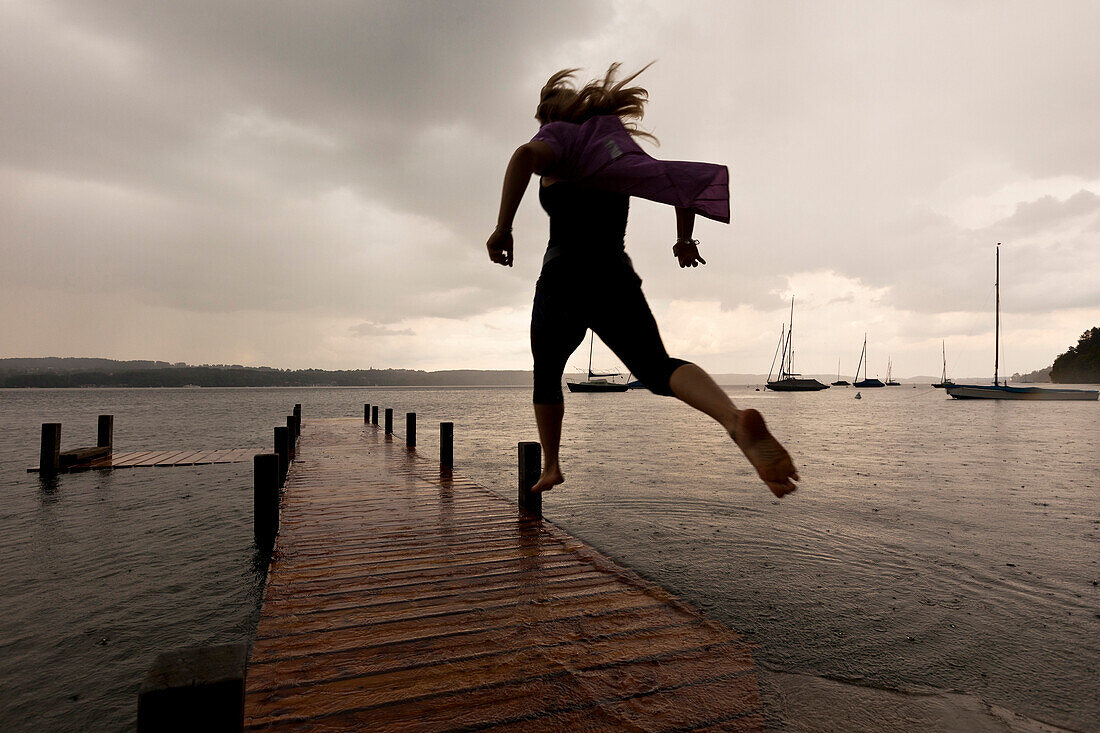 Young woman jumping over pier, high water at Lake Starnberg, Upper Bavaria, Germany, Europe