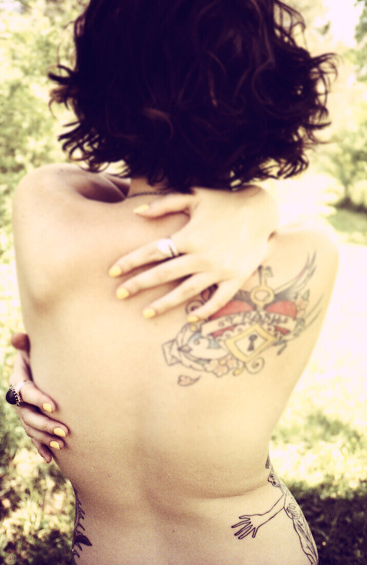 Nackte junge Frau mit Tattoos, die ihre Arme um sich selbst am See schlingt, Rückansicht