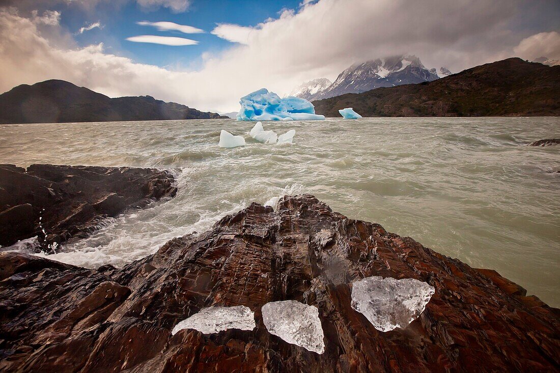 Blue iceberg, rain squalls on Lago Grey, chunks of ice blown onto rocky foreshore, Parque Nacional Torres del Paine, Patagonia, Chile