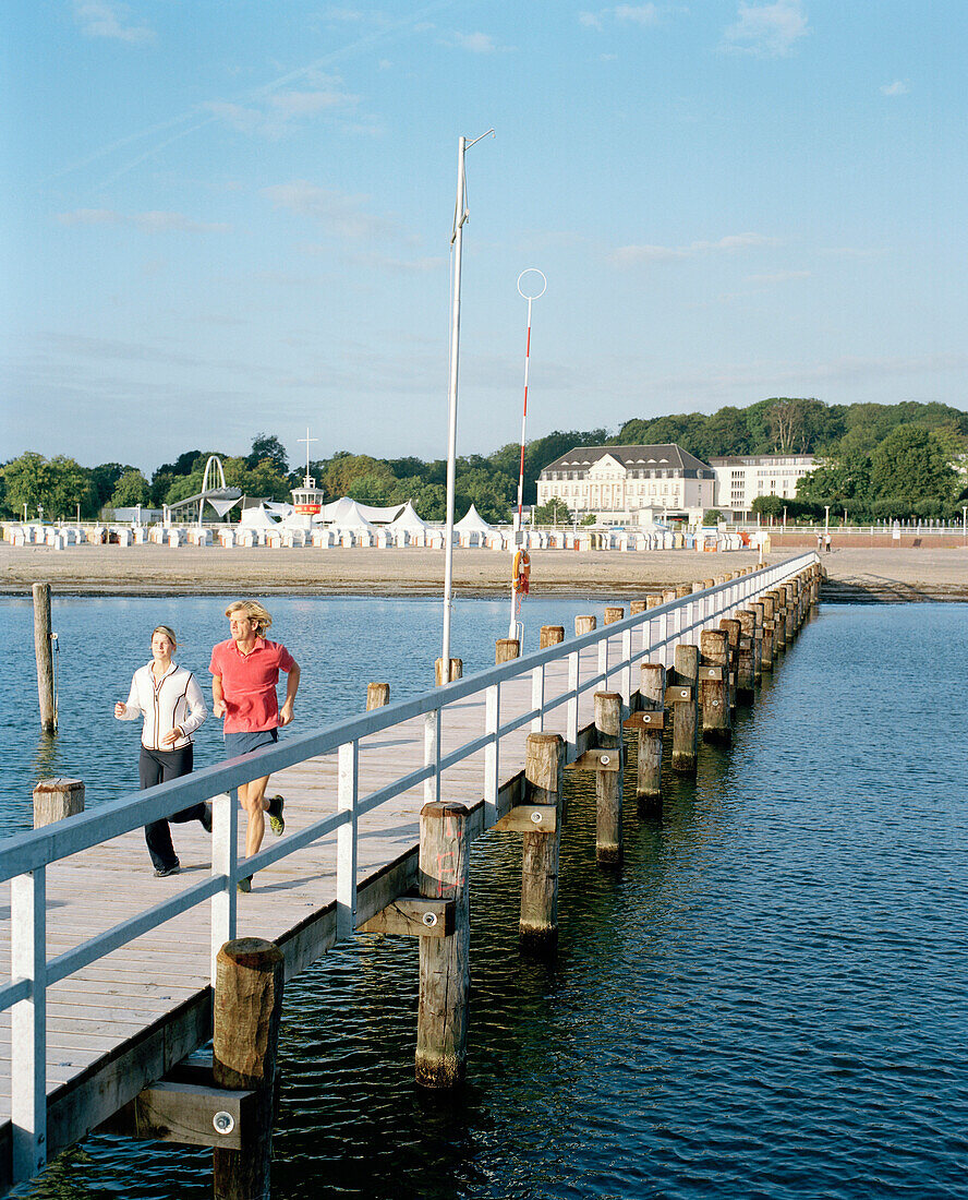 Couple running along a pier at the Baltic Sea, Hotel in background, Travemuende, Luebeck, Schleswig-Holstein, Germany