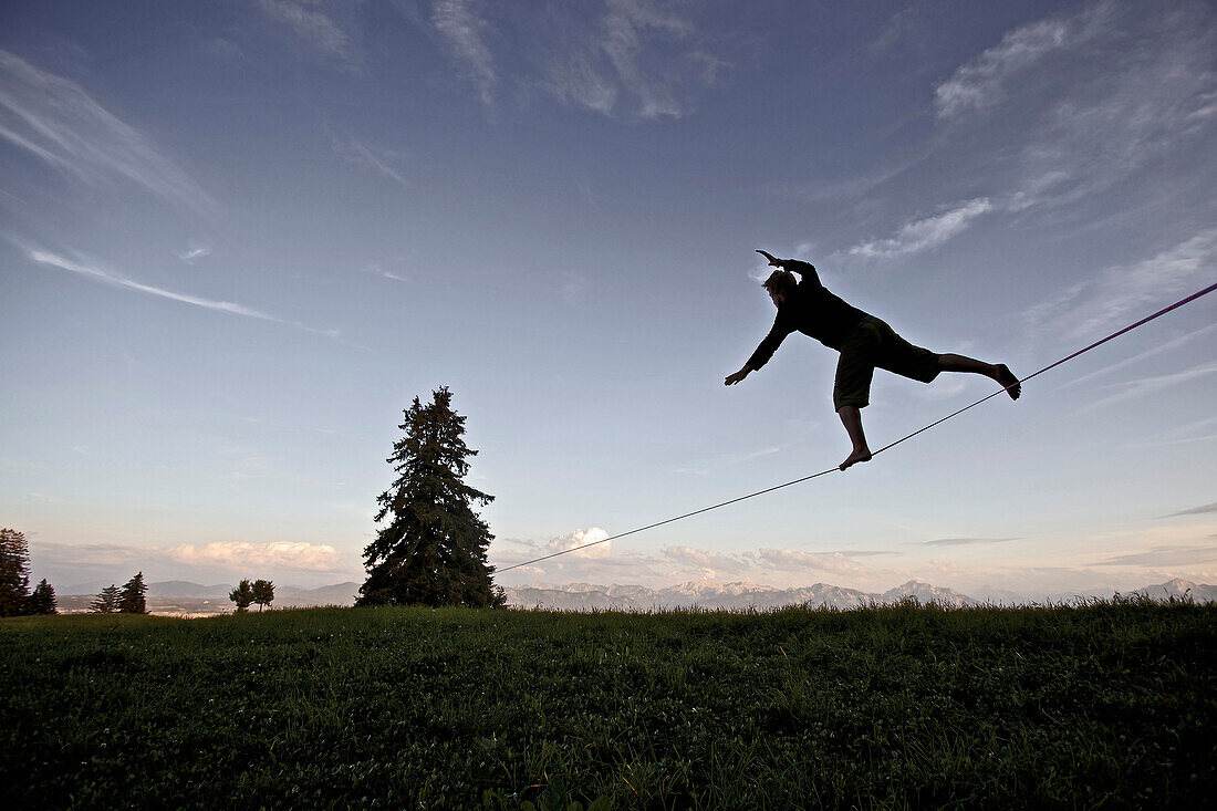Junger Mann balanciert über eine Longline in der Abenddämmerung, Auerberg, Bayern, Deutschland, Europa
