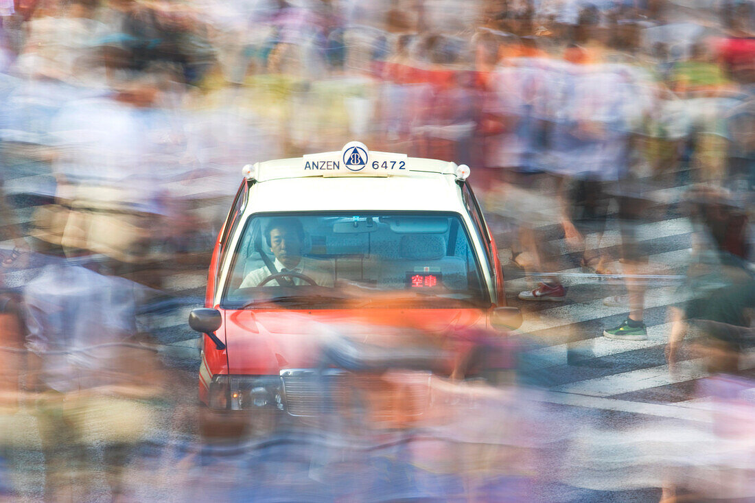 A slow shutter speed blurs a mass of pedestrians that swirl around a stopped taxi at the scramble crossing in front of Shibuya Station, located in the Shibuya District of Tokyo, Japan.