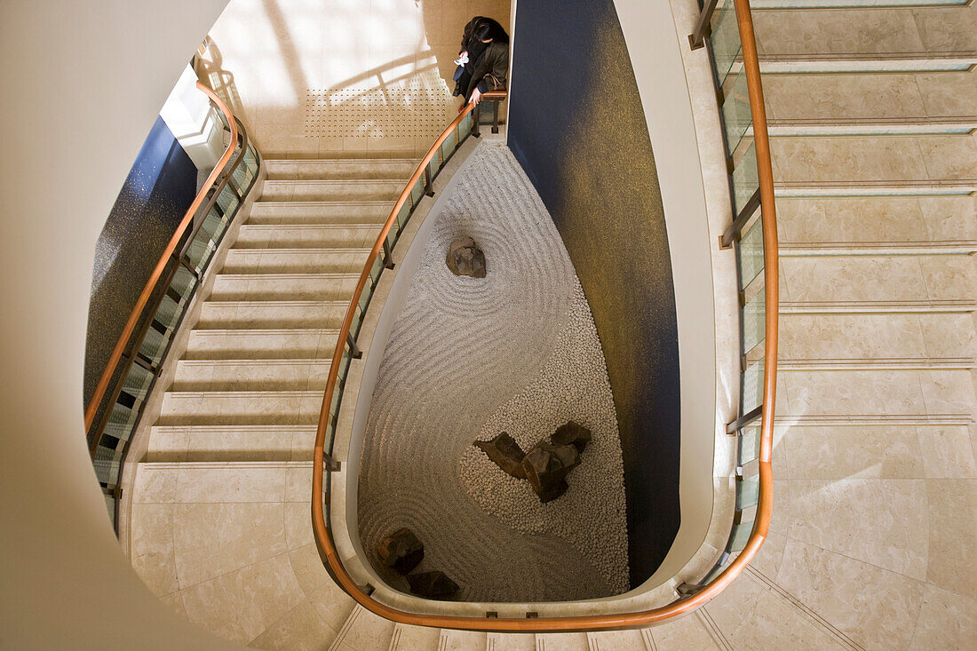 A Japanese woman checks her cellphone at the bottom of a gracefully winding staircase that descends to a small zen-style rock garden inside the Peninsula Tokyo Hotel in the Marunouchi District of Tokyo, Japan.