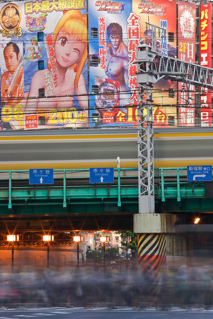 A morning rush of workers with commuter trains overhead cross Yasukuni Dori Avenue with Kabukicho District building-size posters as backdrop at Shinjuku Station in Tokyo, Japan.