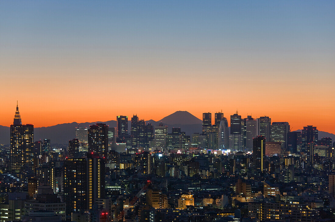 A telephoto view in clear winter twilight captures Mt. Fuji's distinctive peak rising dramatically beyond the skyscrapers (including Tokyo City Hall) and the city lights of the Shinjuku District in Tokyo, Japan.