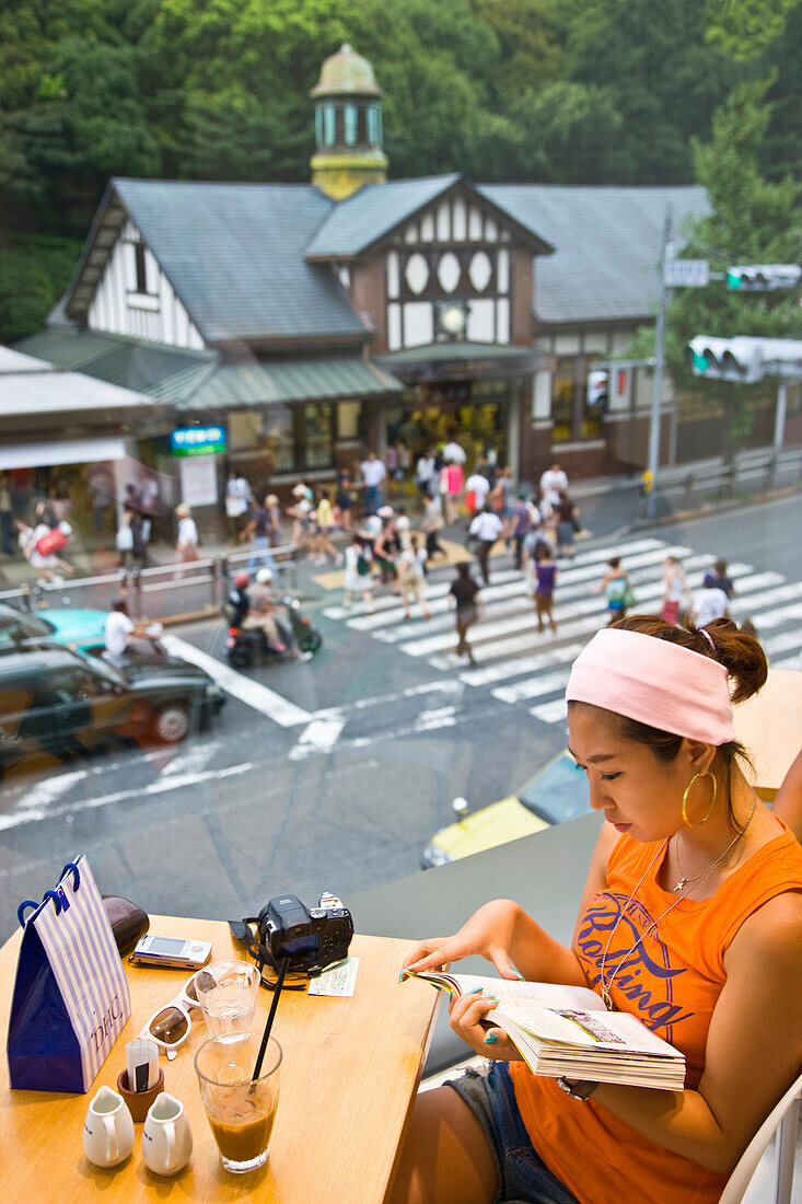 A stylish young Japanese woman relaxes upstairs in a cafe overlooking Harajuku Station and the greenery of Meji Jingu Shrine in the upscale Harajuku District of Tokyo, Japan.