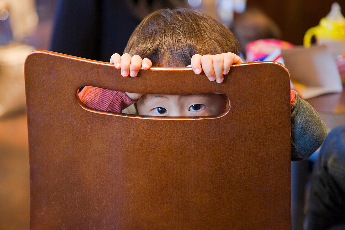 A shyly curious young Japanese boy peers through an opening in the back of his chair at a cafe inside the new Kyoto Station Building in Kyoto, Japan.