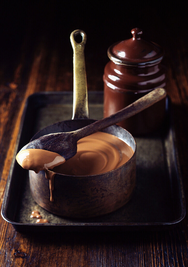 Saucepan of chocolate sauce on tray. Melted Chocolate Sauce in a Panwith a wooden spoon and saucedripping in a tin pan on a rustic wooden surface
