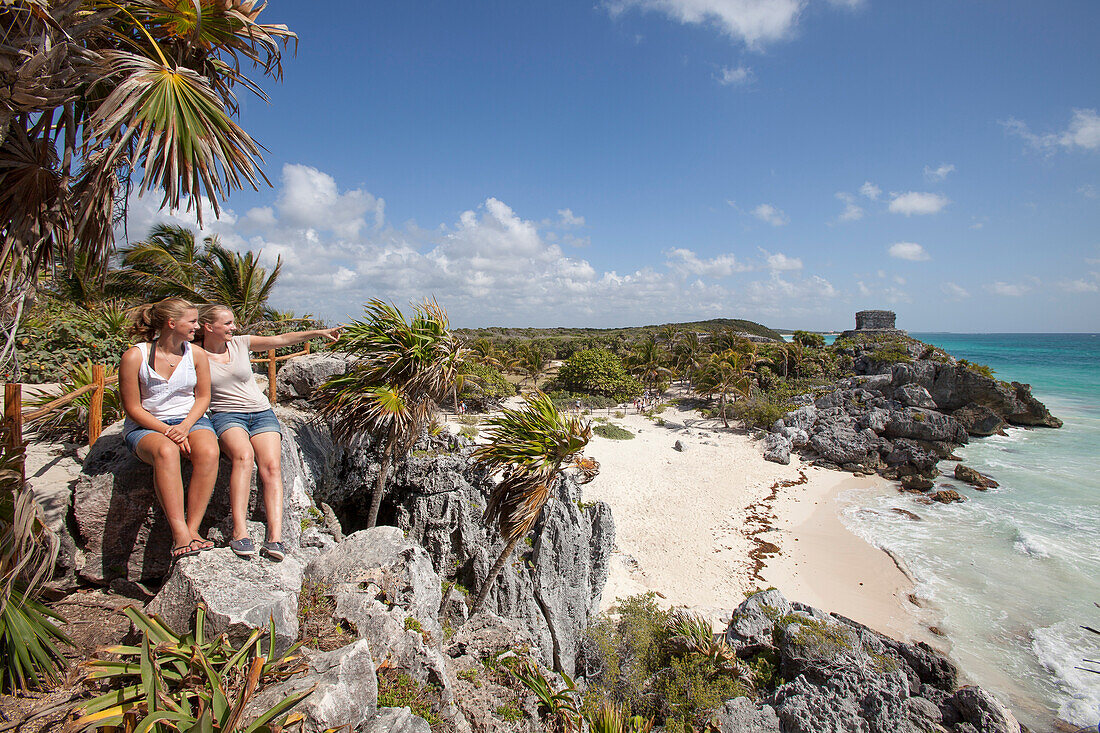 Two young girls and an iguana relaxing on a rock overlooking the beach, ancient Mayan buildings at the Tulum Ruins in the background, Tulum, Riviera Maya, Quintana Roo, Mexico