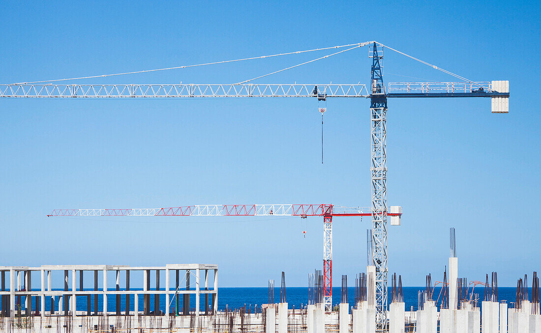 Two cranes at the construction site of a multi-unit building near the coast in Spain. Concrete pillars. Economic development, and economic collapse. Property speculation., Construction Site in Spain