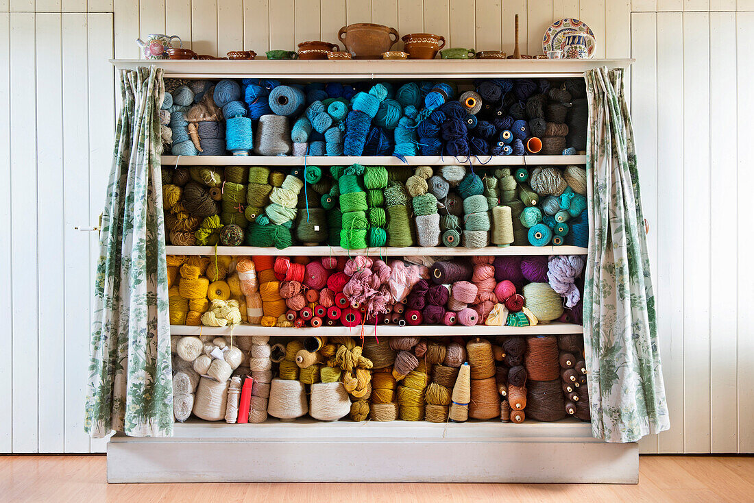 An old yarn or thread cabinet. A set of shelves with curtains, containing wools, yarn, haberdashery, and spools of lint, organised by color. Arranged in a colour pattern., Threads arranged in colour order.