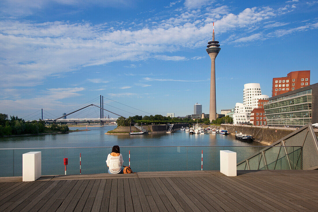 Junge Frau sitzt auf einer Brücke am Medienhafen, Blick auf Rheinturm und Neuen Zollhof, Düsseldorf, Nordrhein-Westfalen, Deutschland, Europa