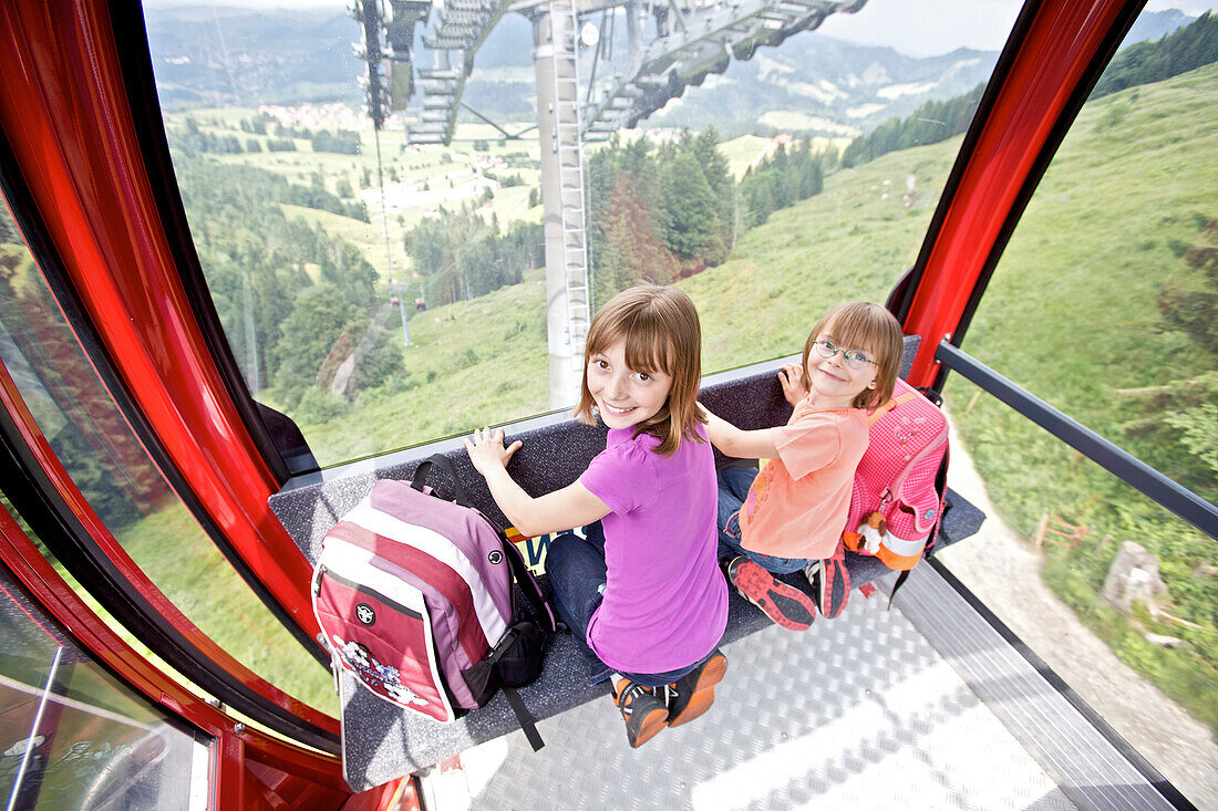 Die zwei Mädchen Hanna and Pia sitzen mit ihren Schultaschen in einer Berggondel, Allgäu, Bayern, Deutschland