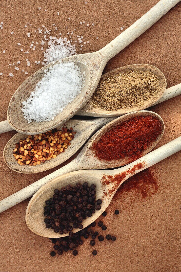 Course Sea Salt, top left, Ground Coriander, top right, Crushed Chillies, middle left, Paprika, middle right and Peppercorns, bottom on wooden spoons on a cork background.