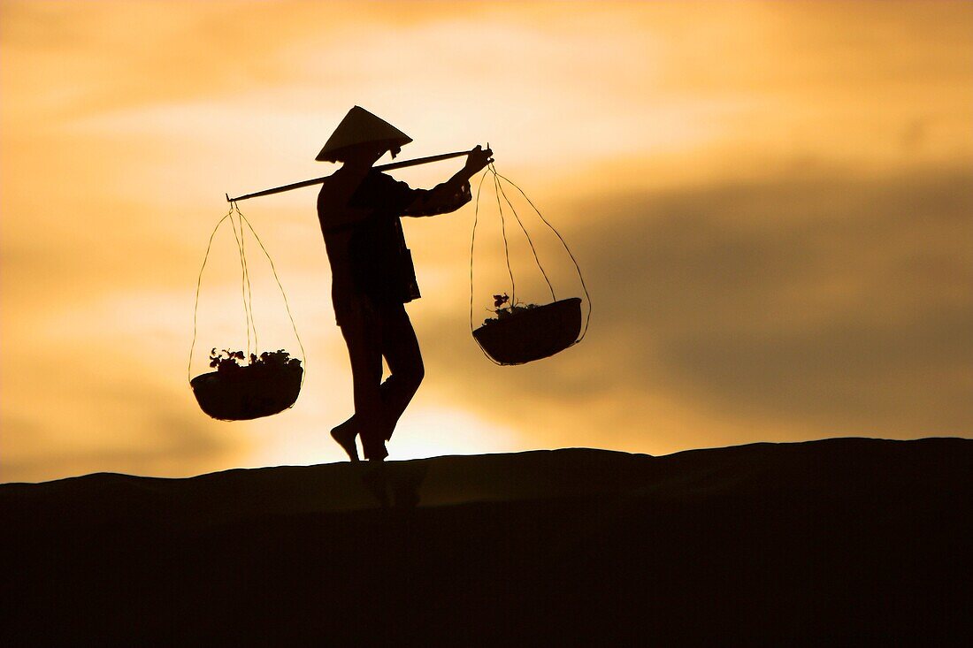 Woman with conical hat carries pannier baskets in sunrise along crest of sand dunes near Mui Ne Vietnam