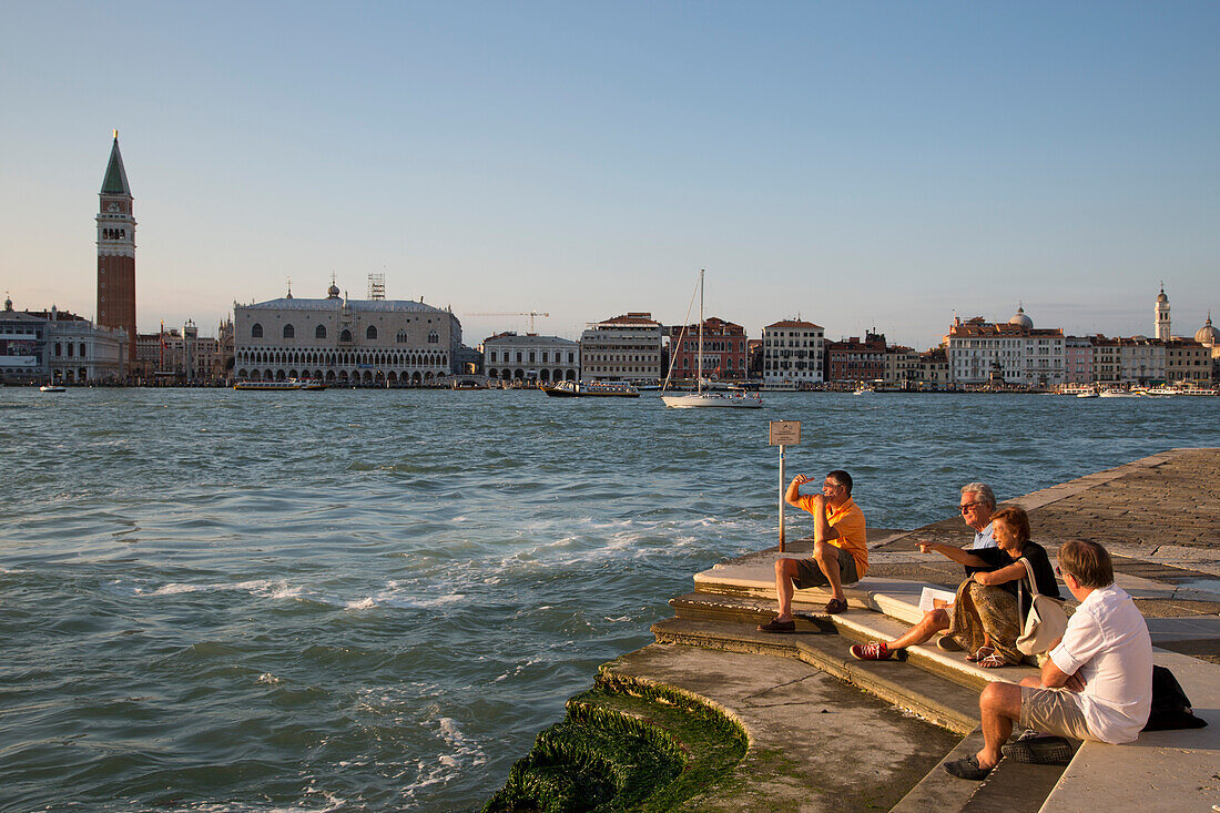 Menschen entspannen sich auf Treppenstufen der Isola di San Giorgio Maggiore Insel mit Blick auf Campanile di San Marco Turm und Dogenpalast am Markusplatz, Venedig, Venetien, Italien, Europa