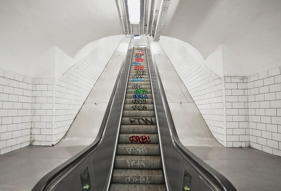 Escalator down to an underground Metro Station in Paris. Transport system. A moving staircase, in a white tiled underground tunnel. sprayed graffiti on stair tread risers.