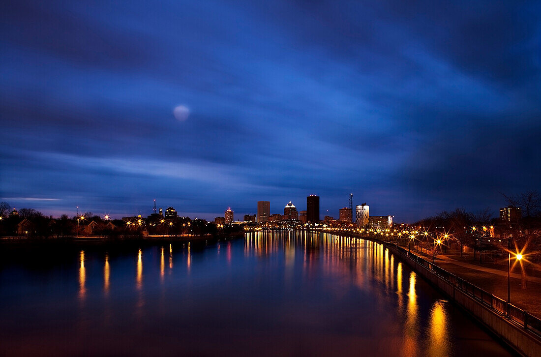 High angle view of the city of Rochester and the Genesee River, at night. Moon in the sky, light clouds. Buildings on the waterfront lit up.