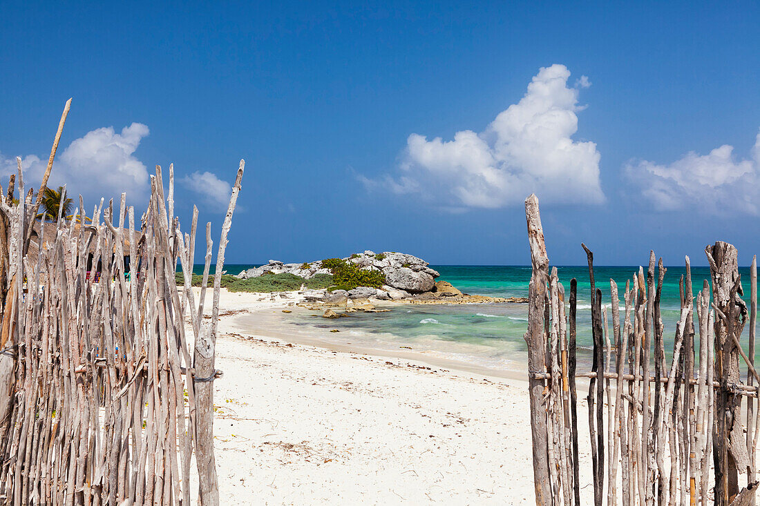 An opening in the fence onto a tropical beach at Tulum. White sand and blue sea. Rocky headland.