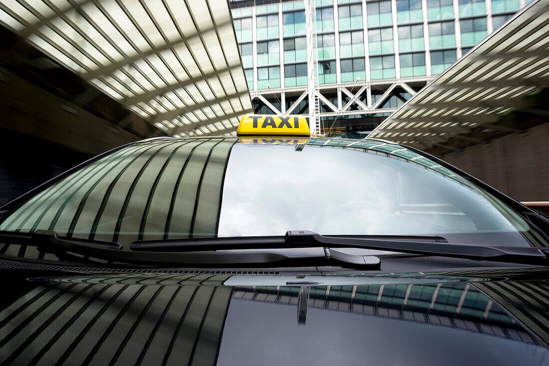 Taxi cab emerging from a tunnel underneath an office building. A front view, with a yellow taxi light on the roof of the car. Reflection on the windscreen of sky and buildings.