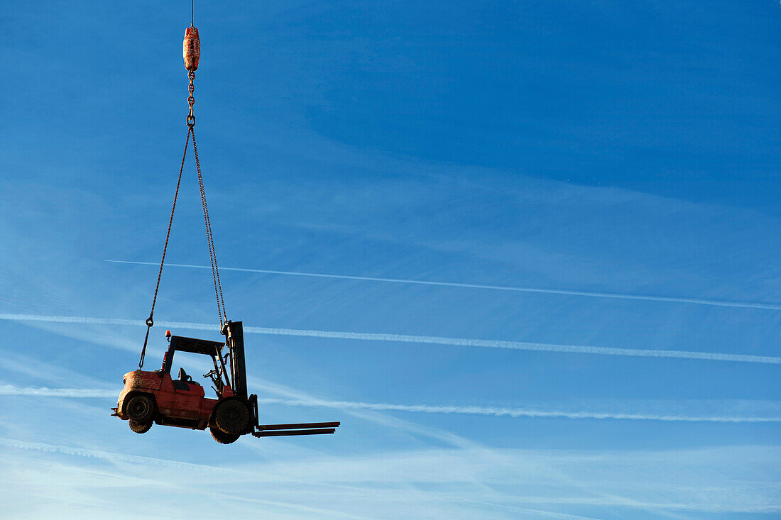 A forklift truck being transported on a platform by a crane. In mid air. View from below. Blue sky.