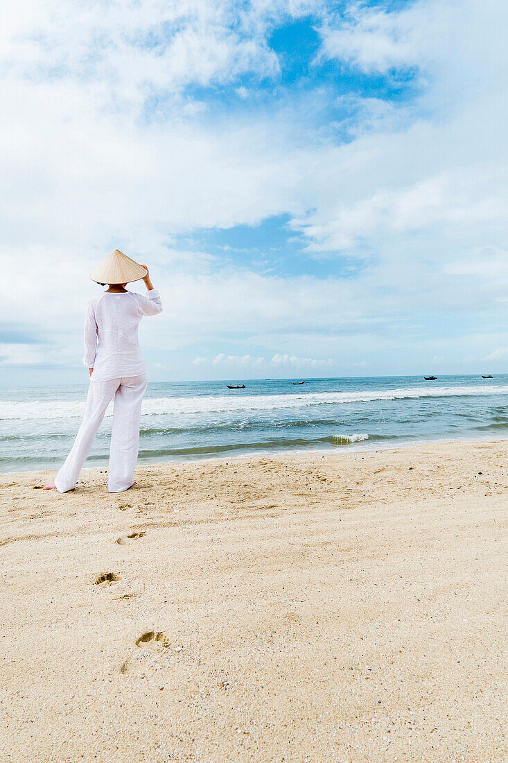 Woman wearing typical clothes and straw hat looking out to sea towards fishing boats, coast of Mui Ne, south Vietnam, Vietnam, Asia