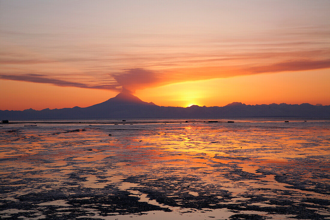 Ash cloud rises from Mt. Redoubt at sunset during low tide near Ninilchik, Alaska