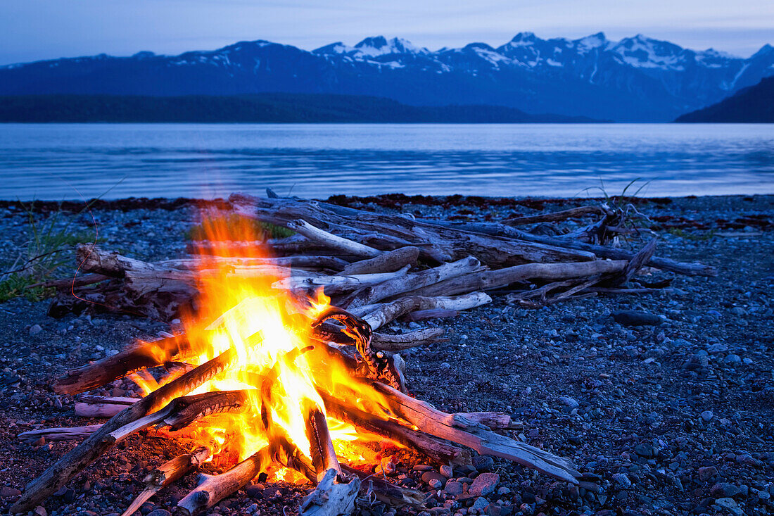 Campfire at dusk on a remote coastal beach in Goose Cove with Fairweather Range in the background, Muir Inlet, Glacier Bay National Park & Preserve, Southeast Alaska, Summer