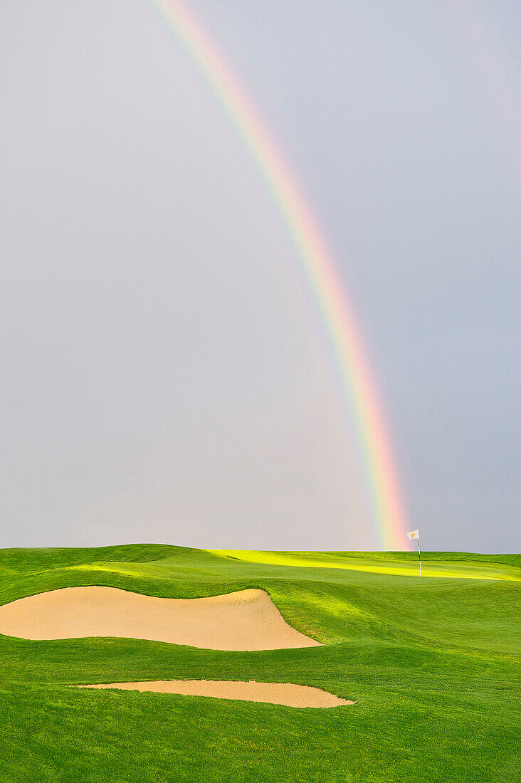 Rainbow and sunbeam shine on putting green and golf pin, vertical photograph