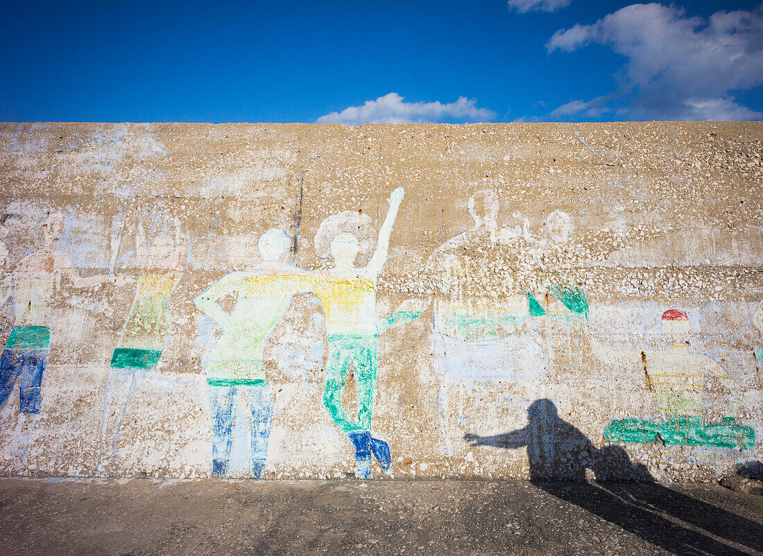 A coastal town.  Marina seawall with graffiti images of people. Shadows of two people on the ground.