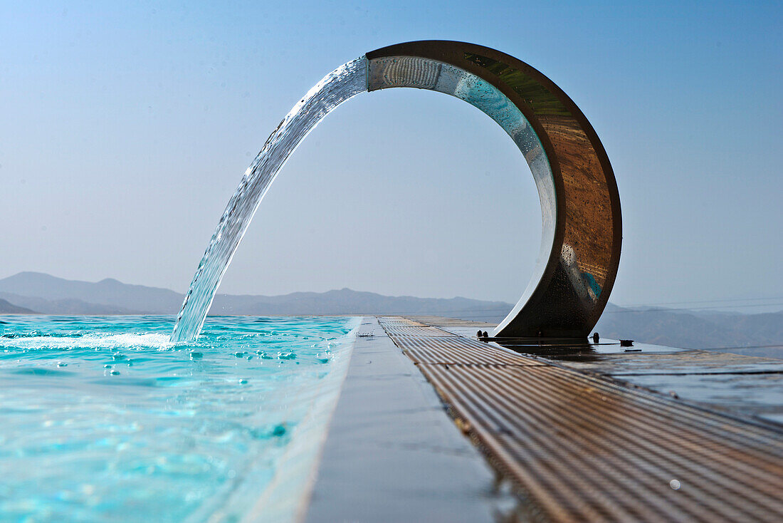 A curved stainless steel water fountain with water flowing into an infinity pool. Hilltop view over mountains.