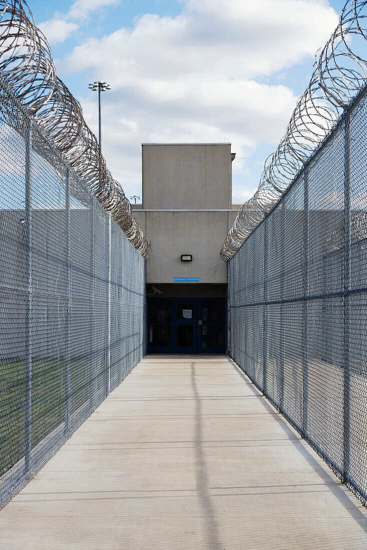 Prison fence and barbed wire at exit of a Correctional Facility. A narrow walkway with high fencing, and coiled razor wire.