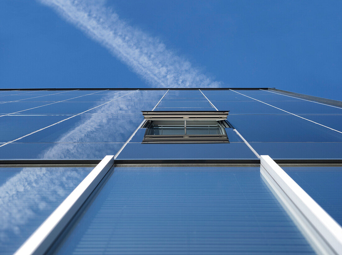 Low angle view of a vapour trail in a blue sky. Large modern building with glass exterior walls. An open window.