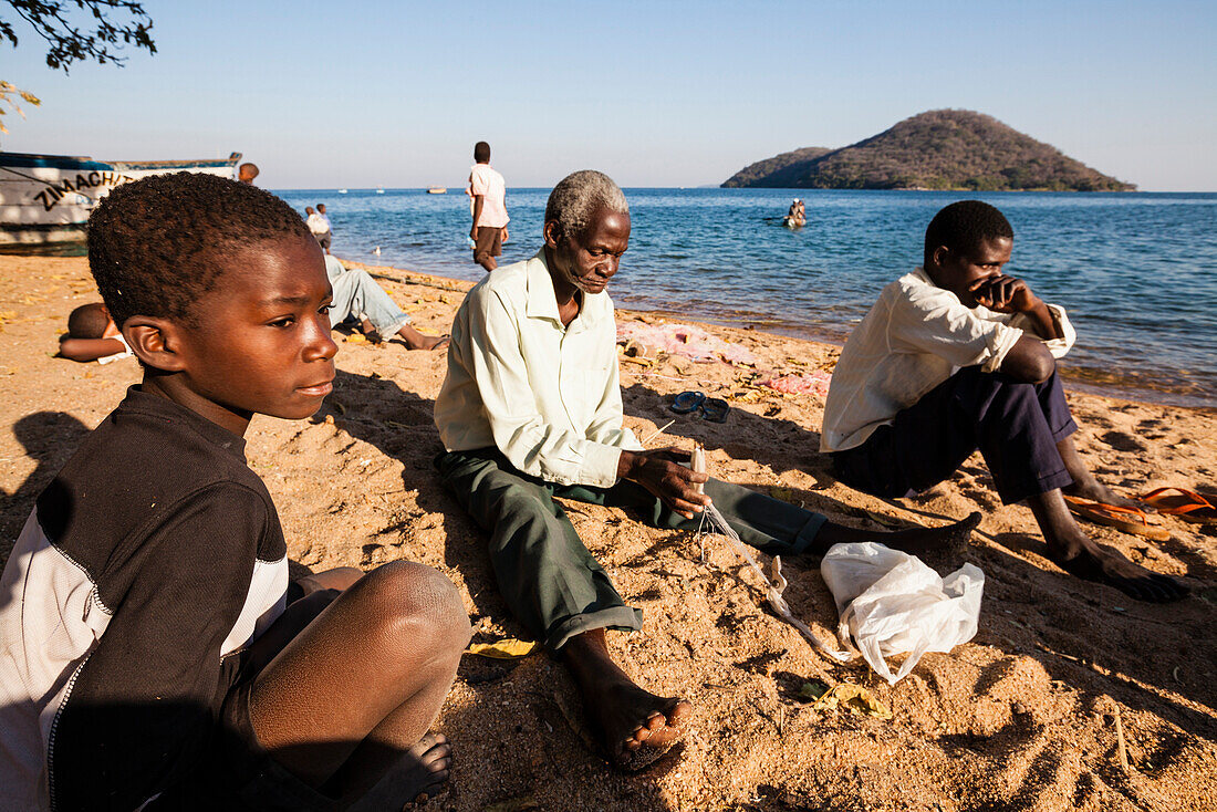 Ein Junge, ein alter und ein junger Mann sitzen am Strand und reparieren ein Fischernetz, Chembe Village, Cape Mclear, Malawi See, Afrika