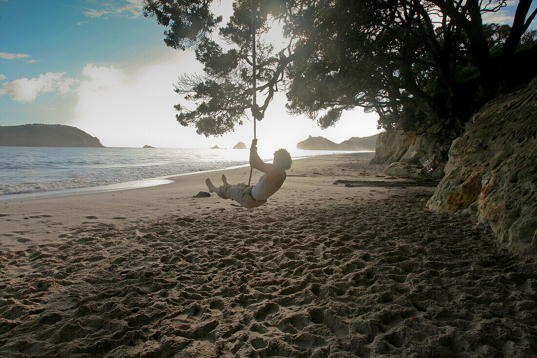 Woman on a rope swing against the sunlight, Hahei Beach Coromandel, North Island, New Zealand
