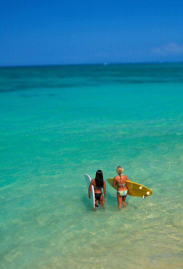 Two females walk into ocean holding surfboards, view from behind, beautiful turquoise ocean out to horizon, blue sky