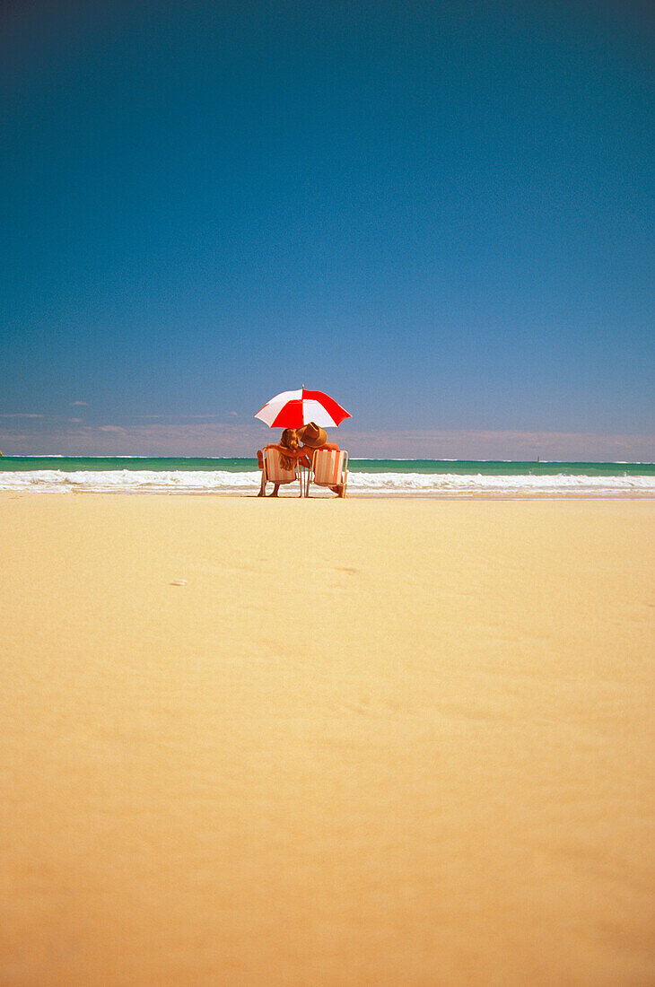 Couple sit under umbrella at beach with white sand and blue sky, view from behind