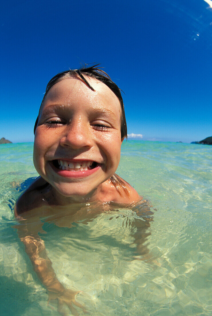 Hawaii, Oahu, Kailua, Extreme close-up of young boy laying in shallow ocean, smiling, cloudless blue sky