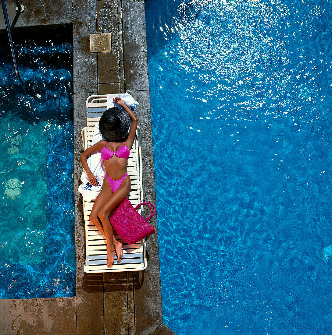 Woman lounging next to swimming pool in pink bikini and black hat over face B1202