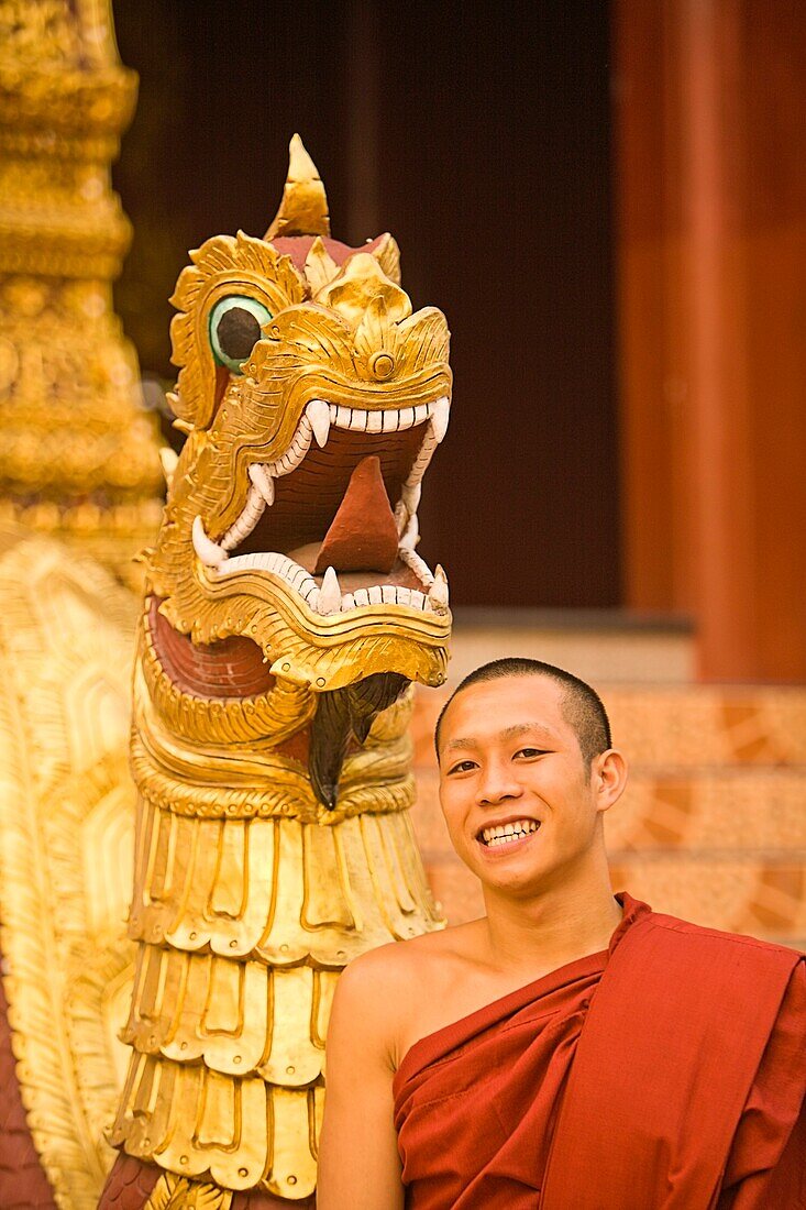 'Young Buddhist Monk Standing In Front Of A Golden Dragon Statue; Chiang Mai, Thailand'